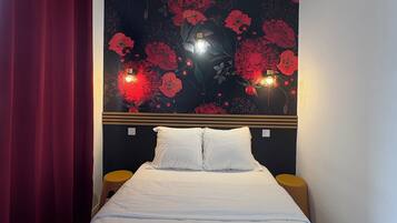 A hotel room with a bed, red curtains, a floral wall decoration, and two bedside lamps.
