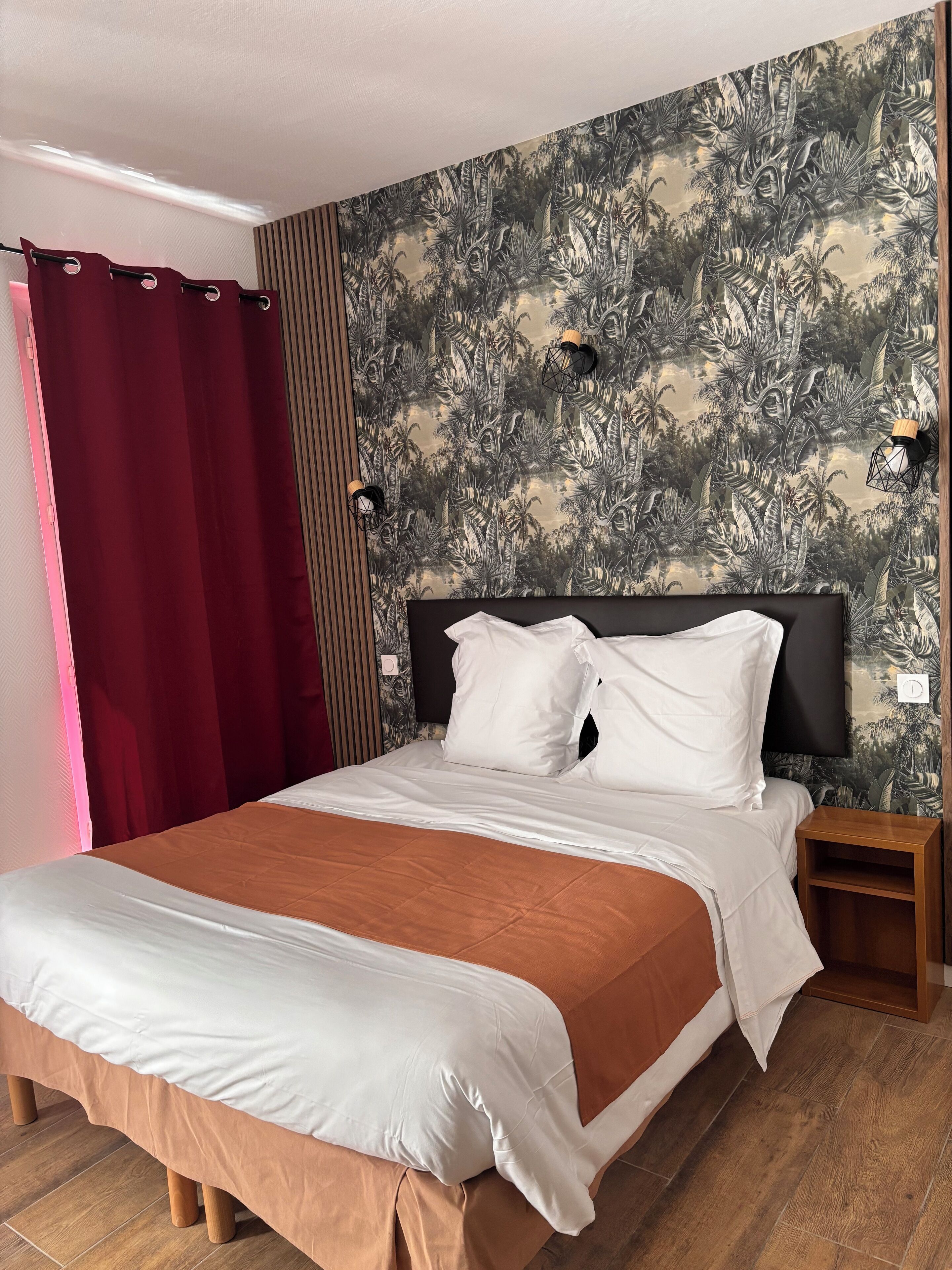 Una cama bien hecha con ropa de cama blanca y marrón, un cabecero oscuro y una mesita de noche de madera con una lámpara. La habitación tiene un papel tapiz tropical y una cortina roja.