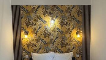 Een net opgemaakt bed met een bruine sprei, twee kussens en een klein nachtkastje met een lamp. De muur achter het bed is voorzien van decoratief behang met een bloemenpatroon en twee wandlampen.