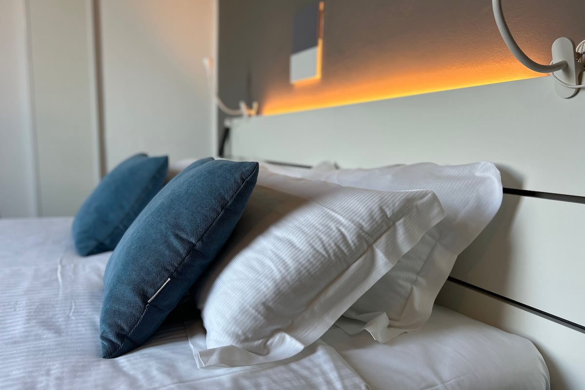 Een opgemaakt bed met witte en blauwe kussens, een wit hoofdbord en een wandlamp.