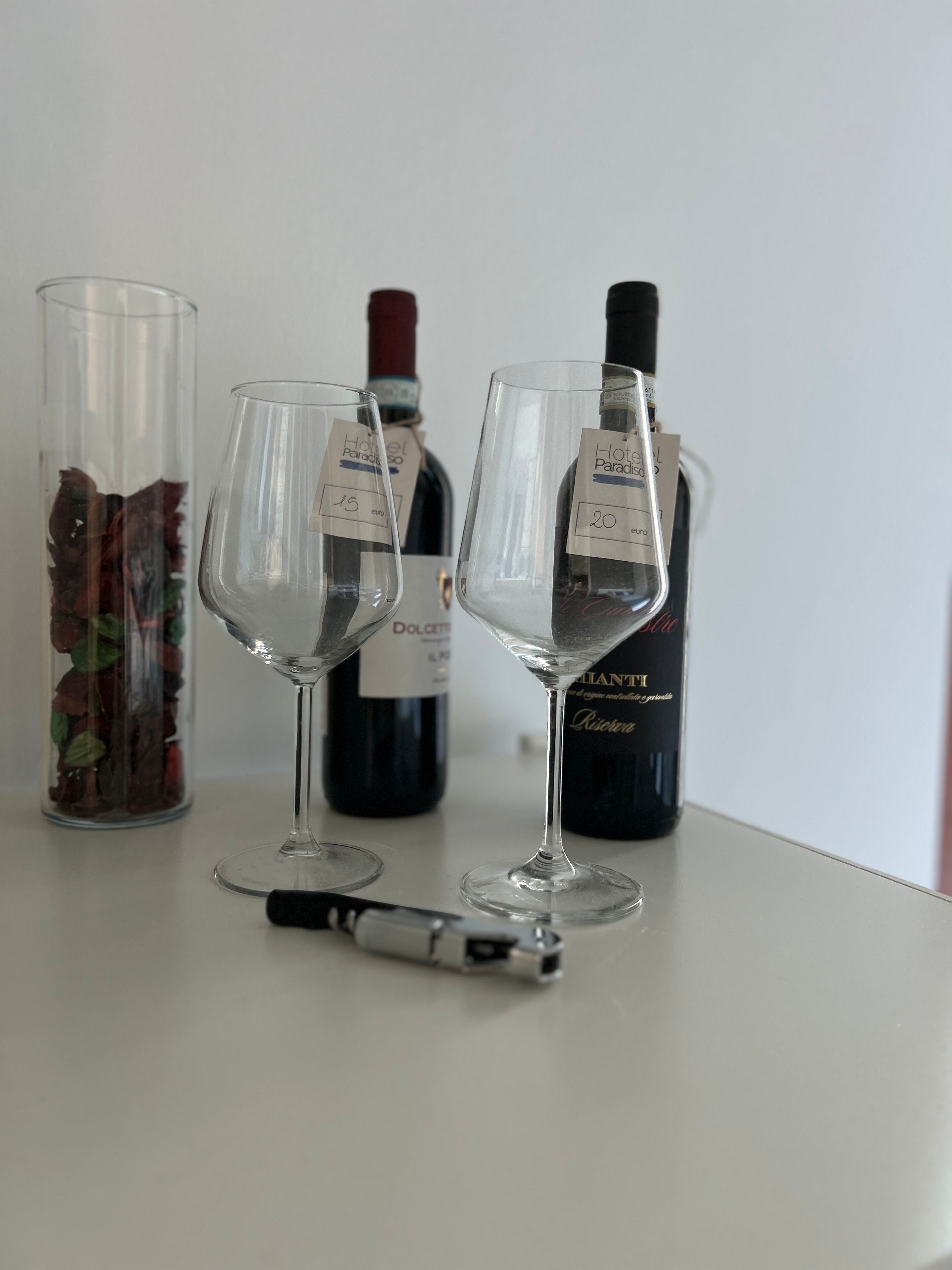 Two wine glasses on a table with a wine bottle and a glass container of dried flowers.
