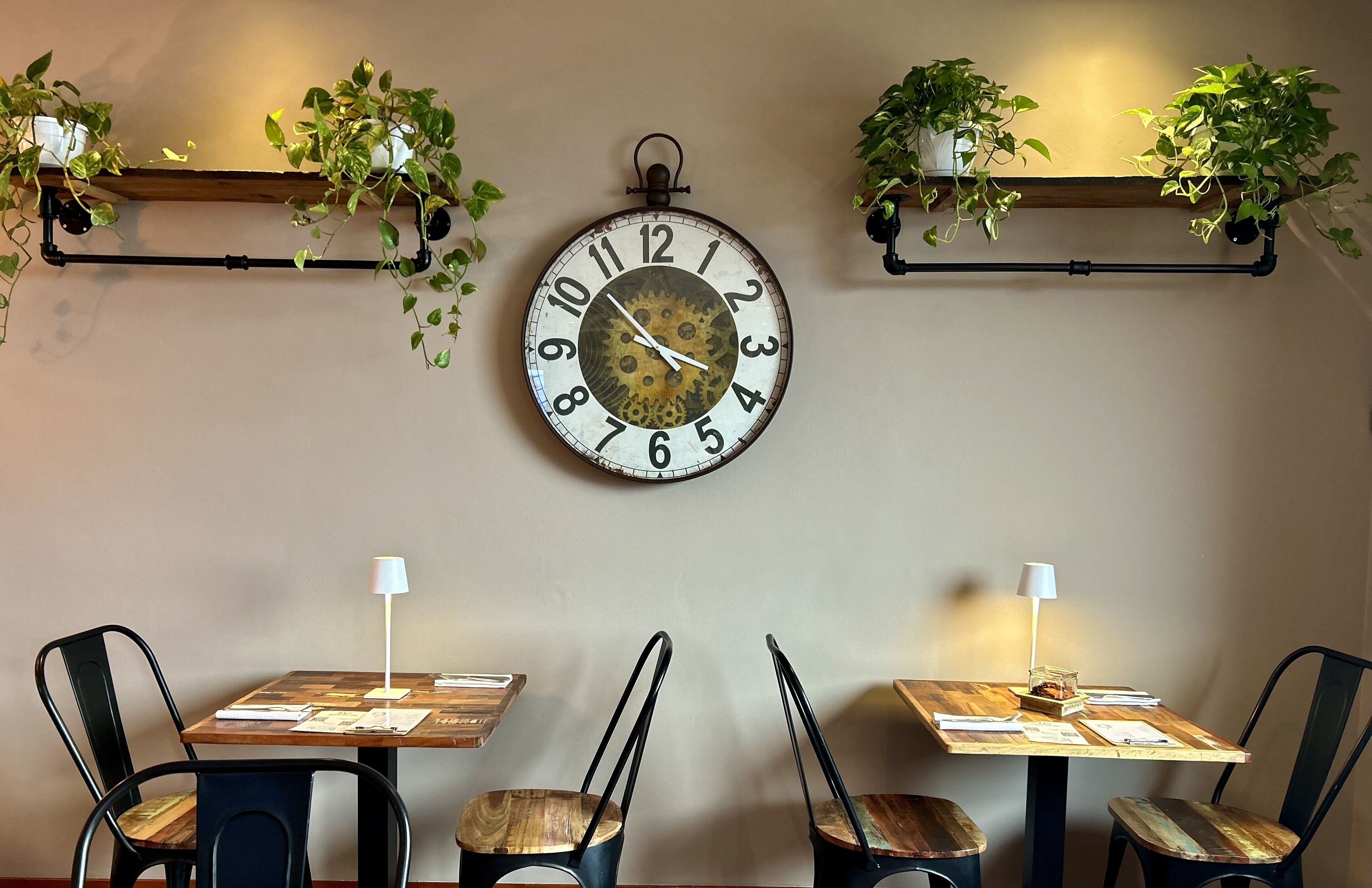 A dining area with a wall-mounted clock, wooden tables, black chairs, and potted plants.