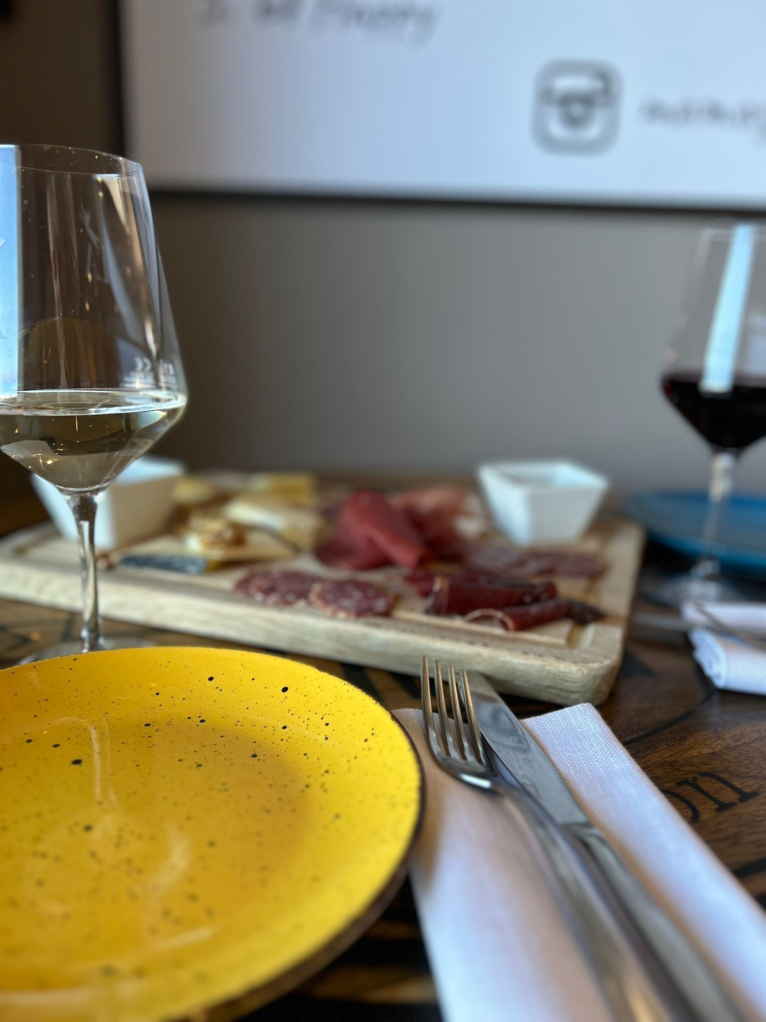 A dining table with a yellow plate, a glass of white wine, and a wooden board with various meats.