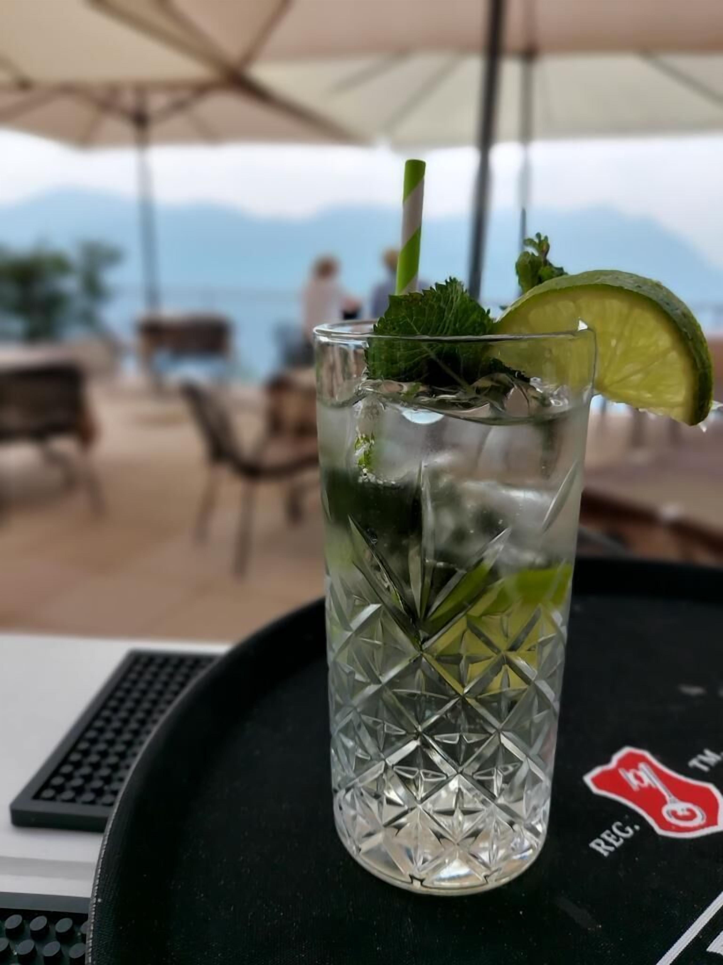A glass of iced drink with a lime wedge and mint on a tray with a logo, set outdoors with a view of mountains.