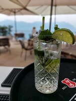 Un vaso de bebida helada con un trozo de lima y hojas de menta sobre una bandeja con un logotipo, todo en un espacio al aire libre con vistas a montañas.