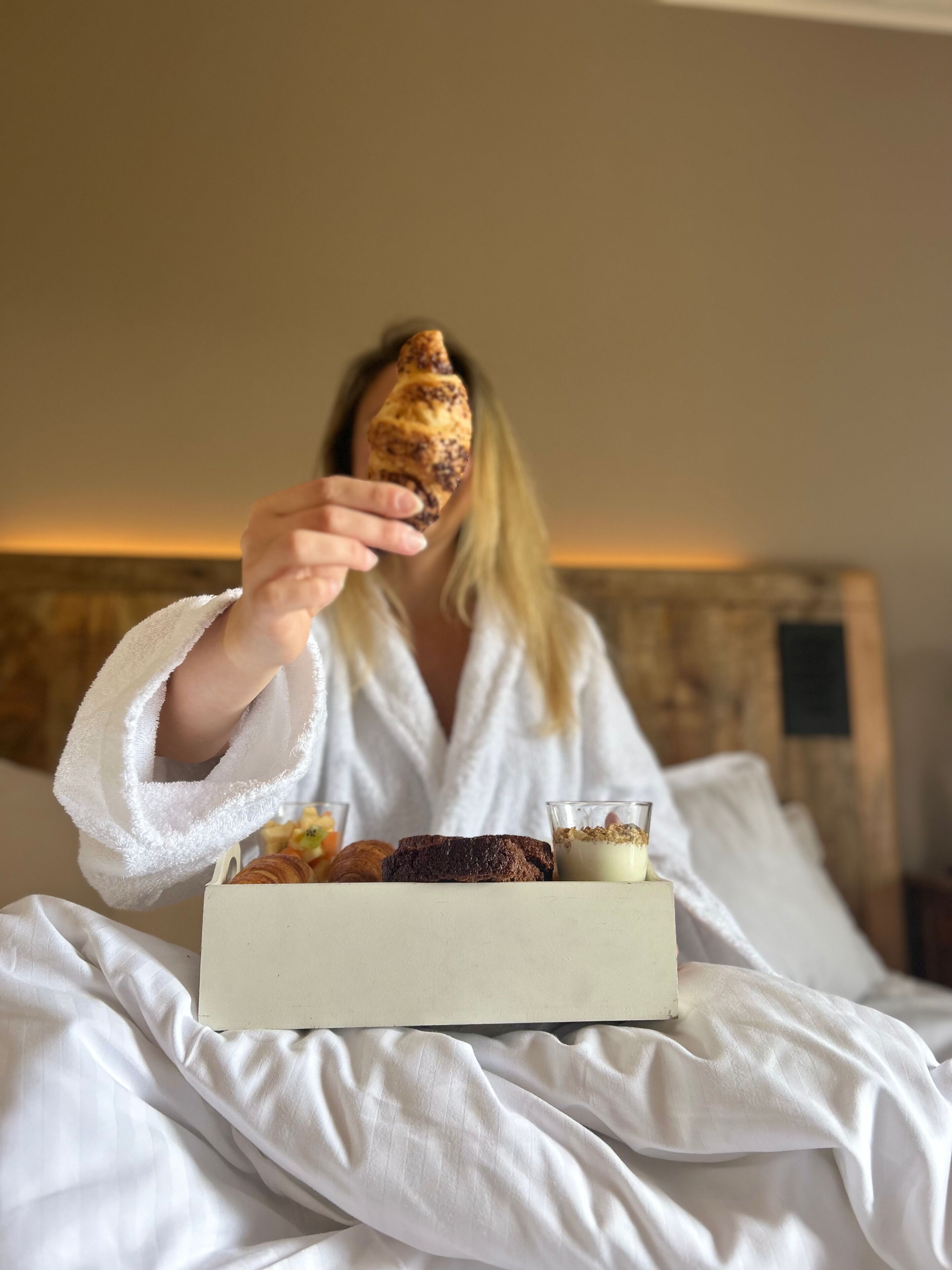 Uma pessoa vestindo um roupão branco sentada em uma cama, com uma caixa de doces e um copo de iogurte.