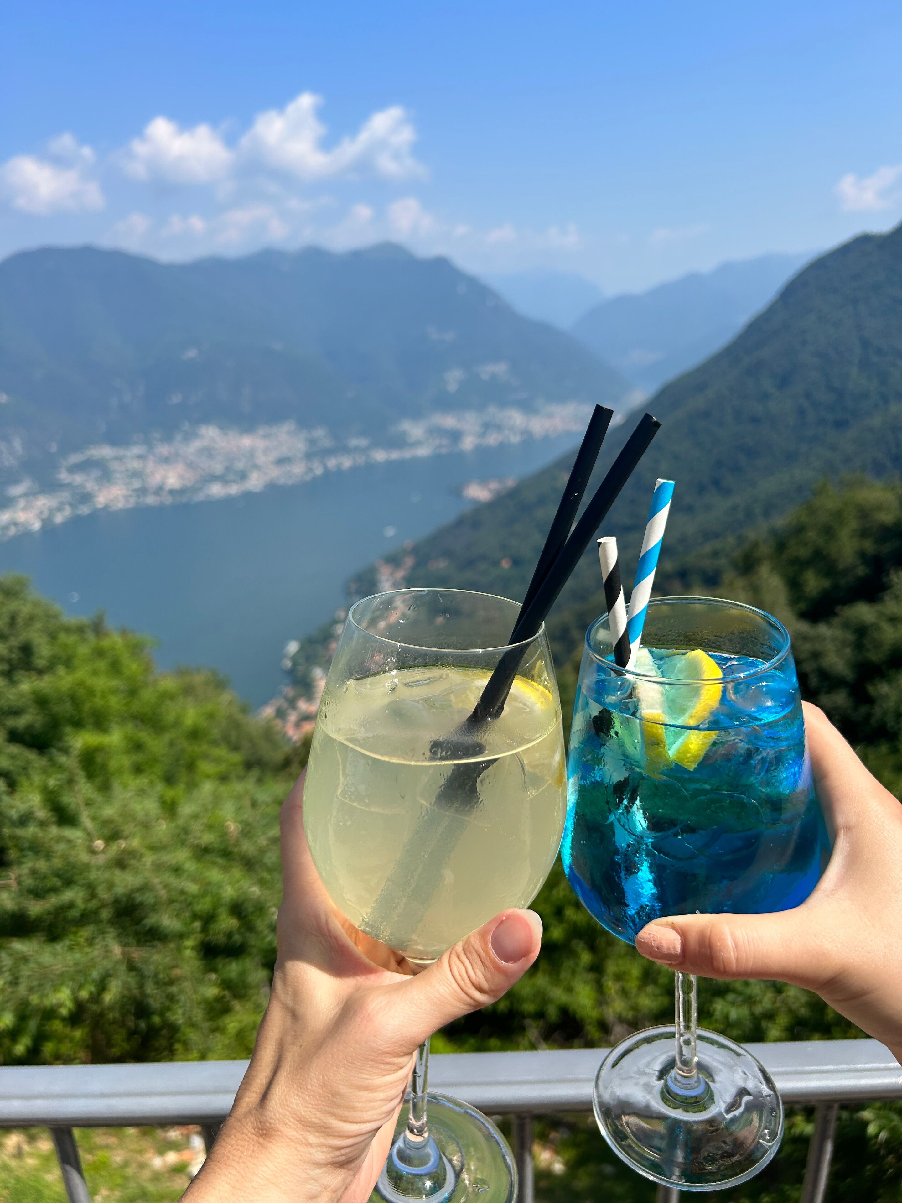 Two hands holding glasses of drinks with straws, one with a lemon slice, against a scenic mountain backdrop.
