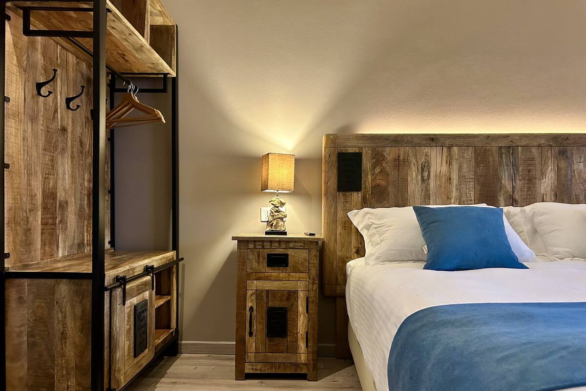 Een houten stapelbed met nachtkastje en lamp, een bed met wit en blauw beddengoed, en een houten hoofdbord.