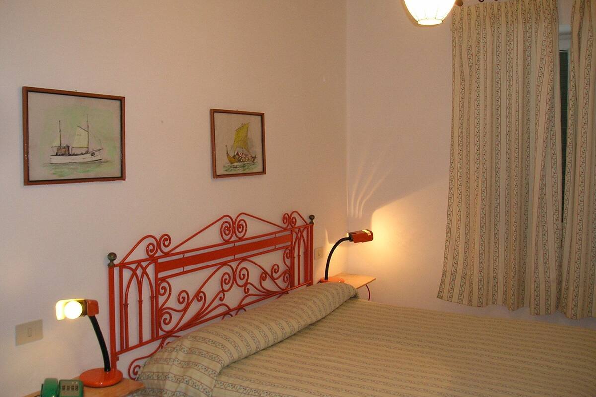Un dormitorio con una cama, cabecera de metal rojo, dos cuadros enmarcados, una lámpara de noche y una ventana con cortinas.