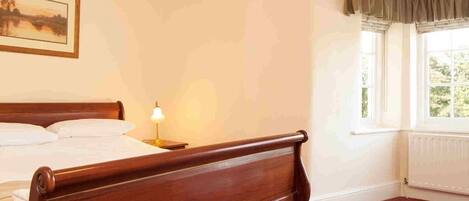A bedroom with a wooden bed, a nightstand with a lamp, a window with curtains, and a painting on the wall.
