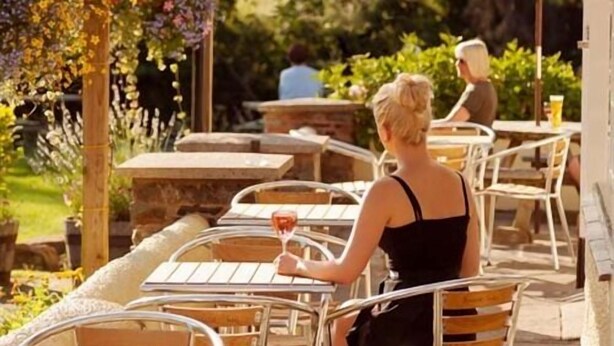 An outdoor patio with metal chairs and tables, a wooden pergola with flowers, and a person holding a glass. - Anchor Inn (Dulverton)