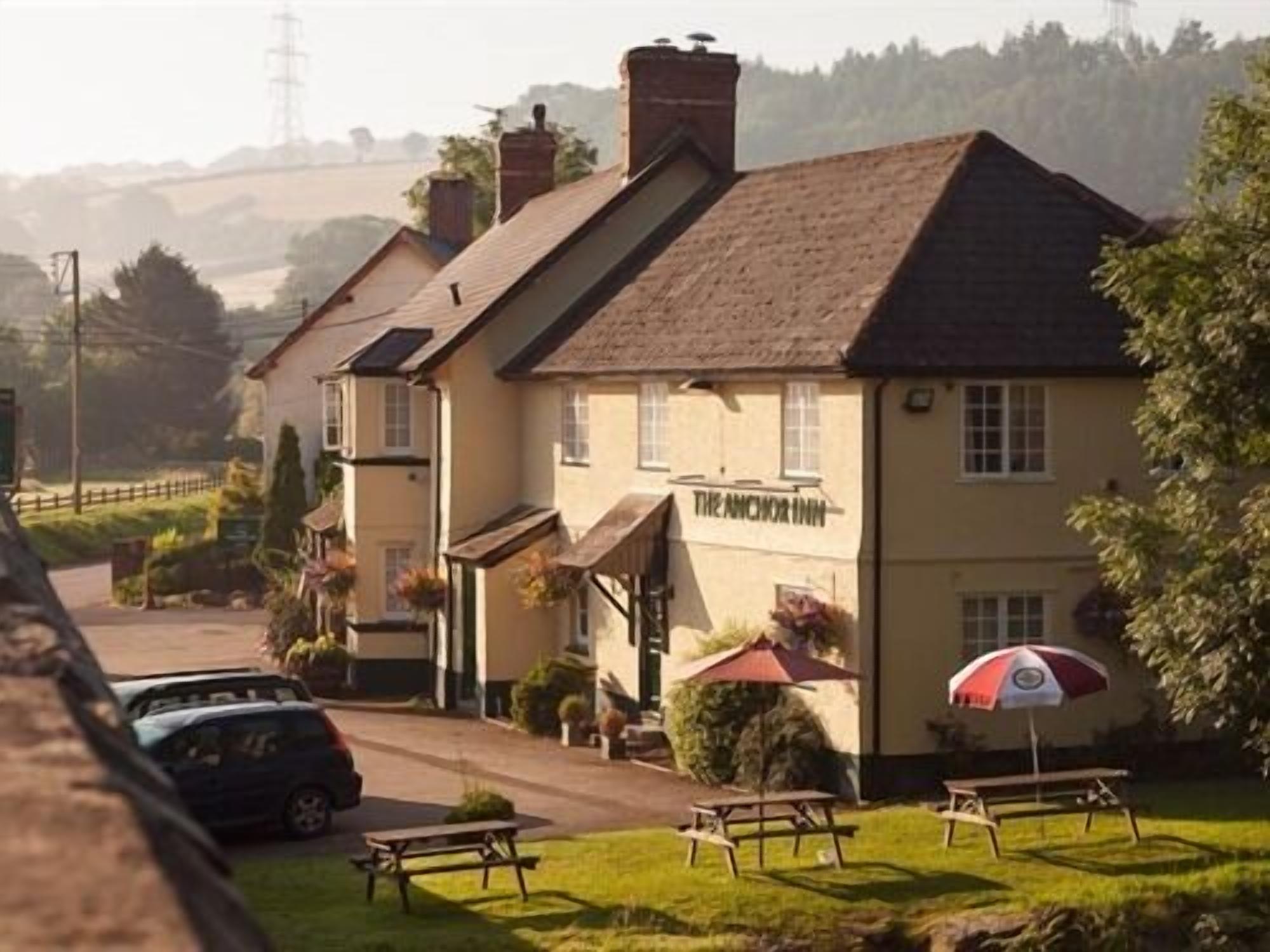 A traditional pub named "The Anchor Inn" with a classic architectural style, featuring a prominent entrance, outdoor seating area with tables and umbrellas, and a parking area visible in the foreground.
