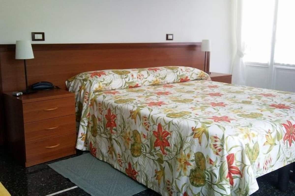Habitación de hotel con cabecera de madera, cubrecama floral, mesita de noche con lámpara y teléfono.