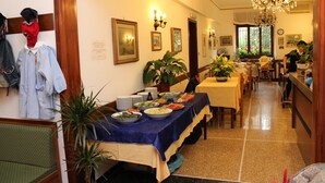 Uma sala de jantar com uma mesa comprida coberta por um pano de mesa azul, cercada por cadeiras. Há plantas em vasos e quadros nas paredes.