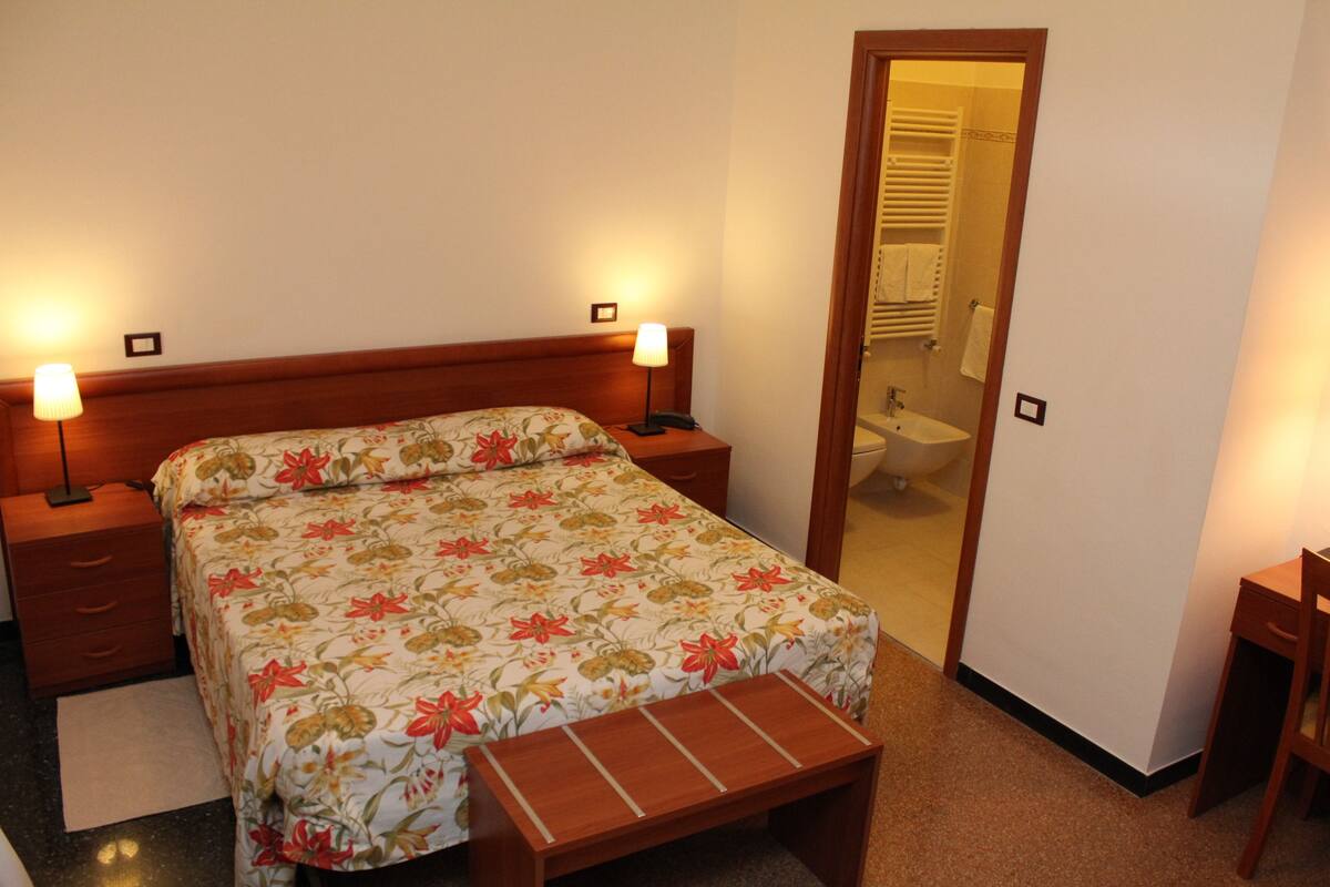 Un dormitorio con cama, mesitas de noche, un banco de madera, un espejo y un baño visible a través de una puerta abierta.