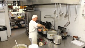 Uma cozinha profissional com eletrodomésticos de aço inoxidável, panelas e frigideiras na bancada, e um chef preparando alimentos.