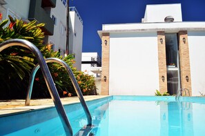 A swimming pool with metal ladders, surrounded by plants and a white building with brick accents. - Pousada Omnimare (Ubatuba)