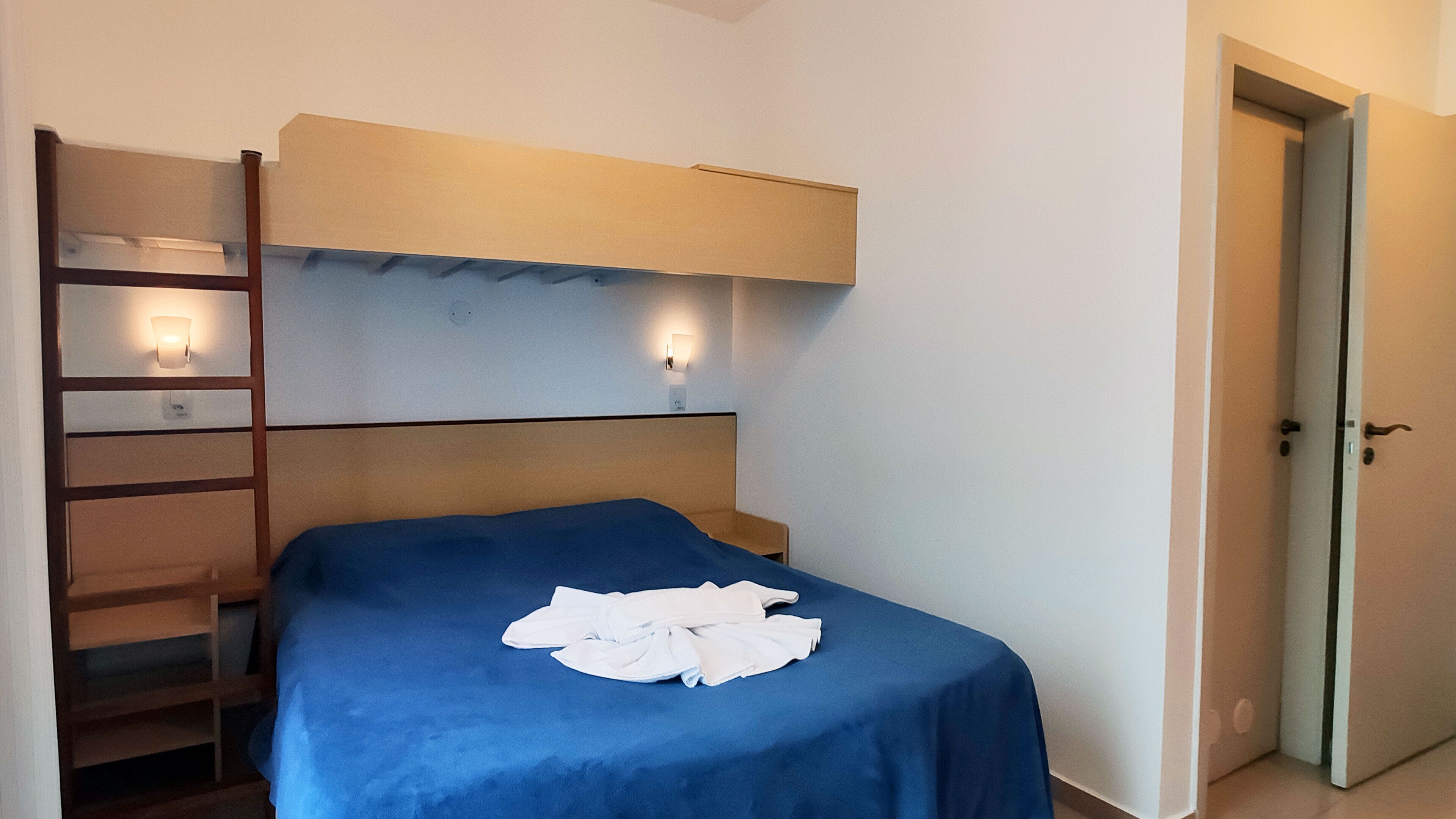 A bedroom with a bed, a nightstand, a wall-mounted shelf, and a door.