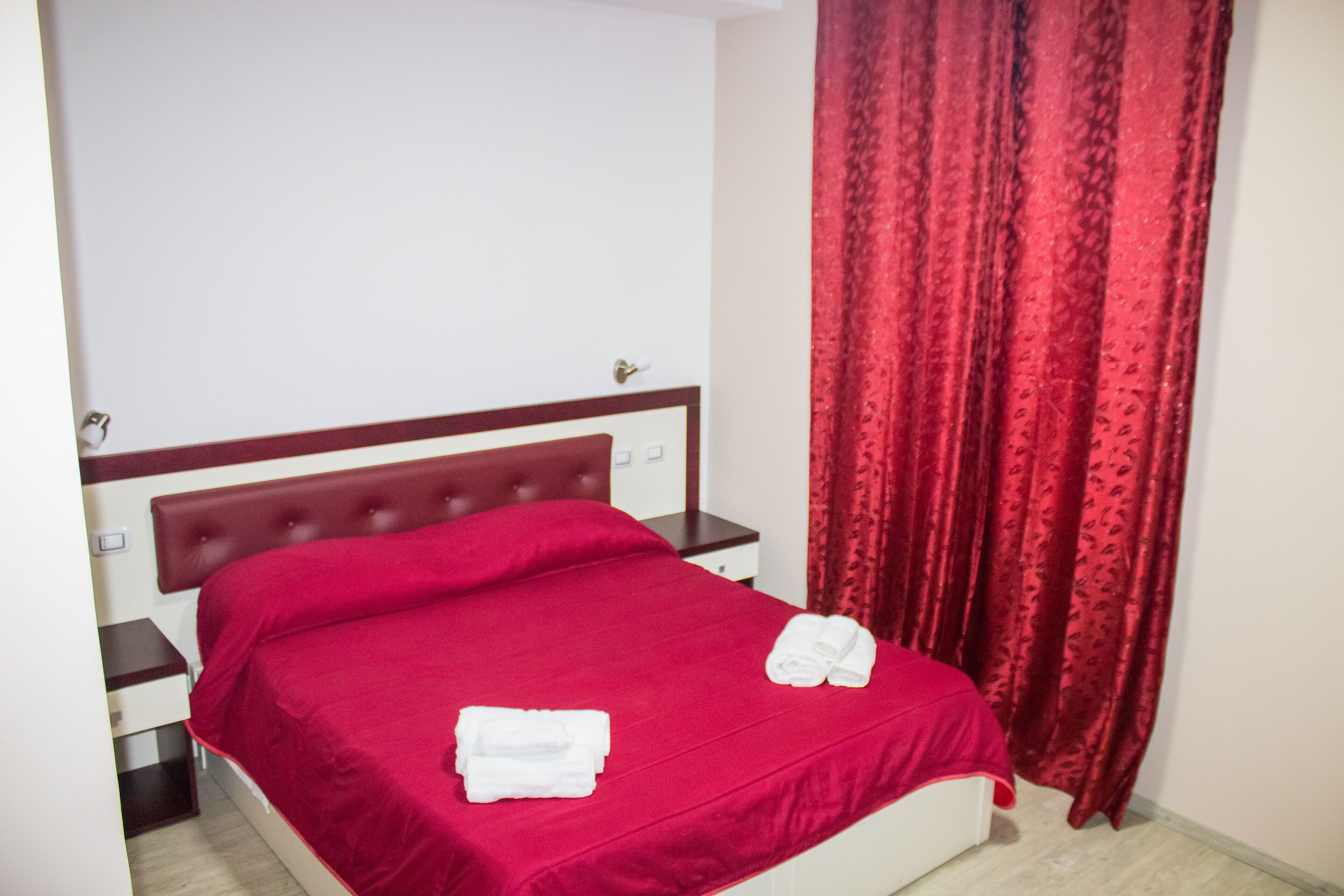 Una cama bien hecha con ropa de cama roja y toallas blancas, cabecera de madera y cortina roja.