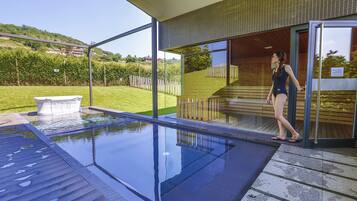A modern outdoor swimming pool with clear glass walls, a wooden deck, and a view of a green landscape.
