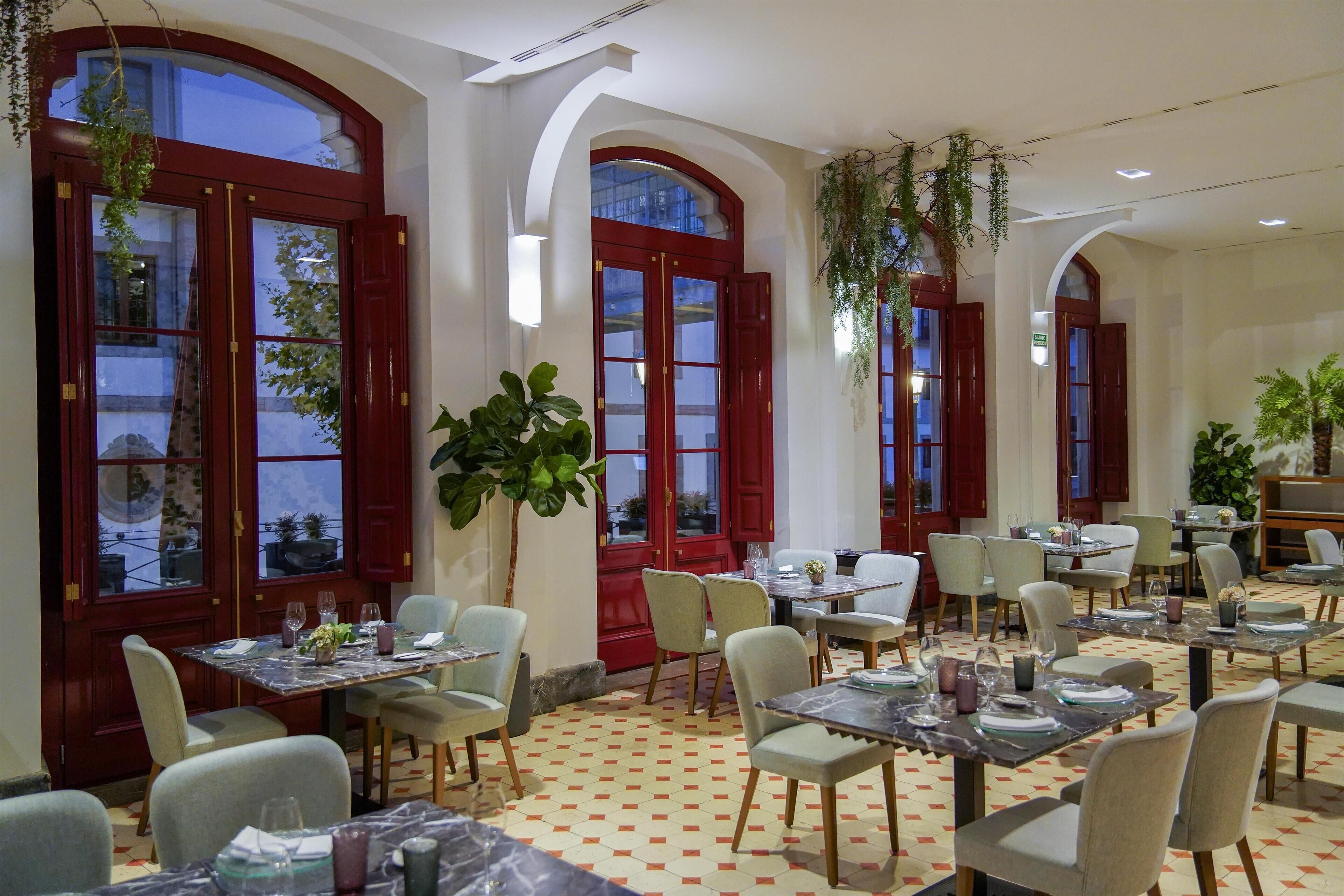 Restaurante com portas vermelhas, paredes brancas e chão de azulejos. Mesas montadas com cadeiras e pratos.