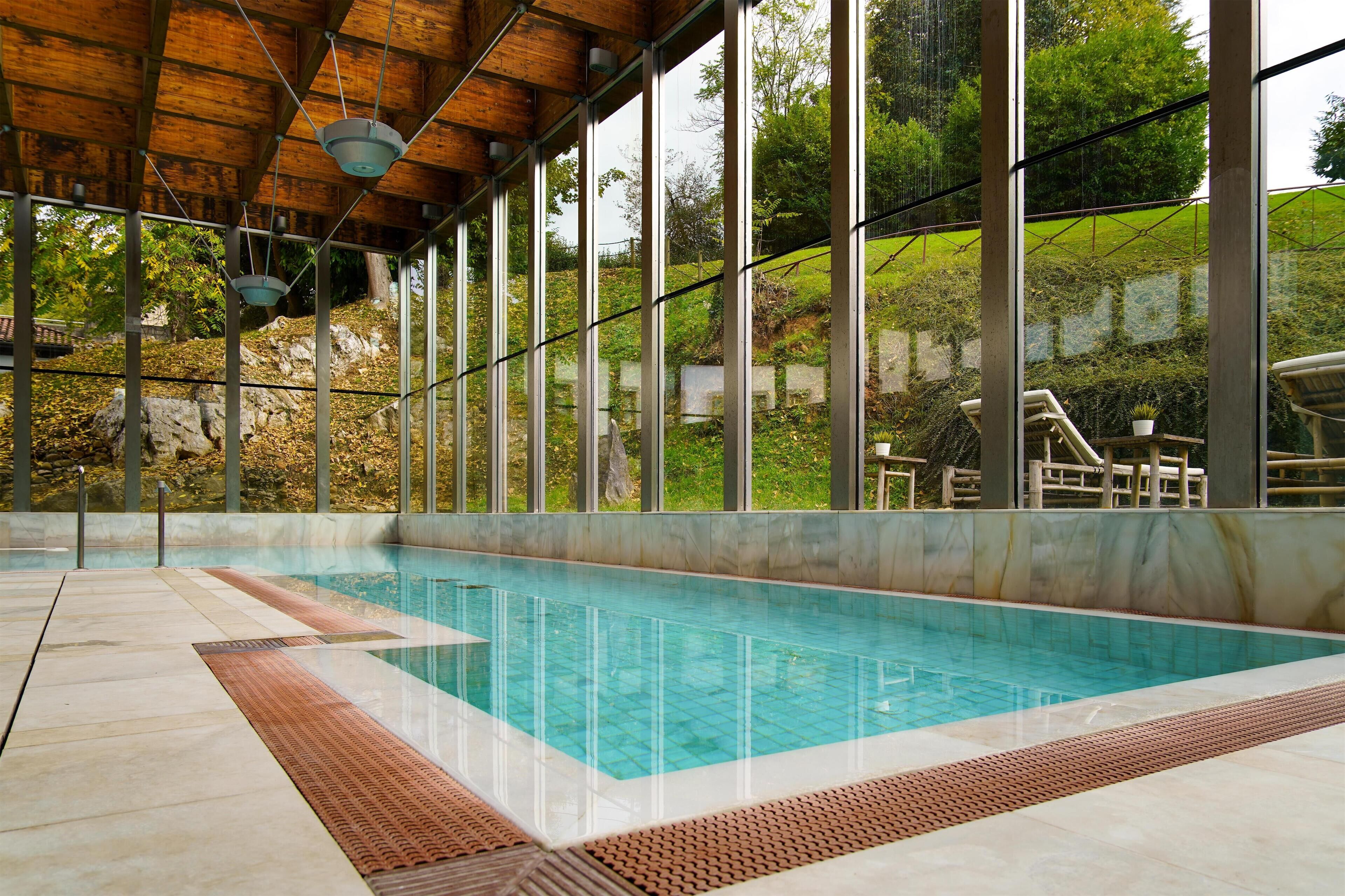 An indoor swimming pool with a wooden ceiling and large windows overlooking a landscaped area.