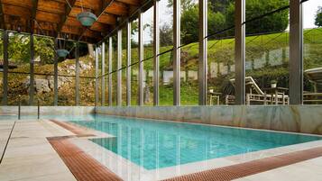 An indoor swimming pool with a wooden ceiling and large windows overlooking a landscaped area.