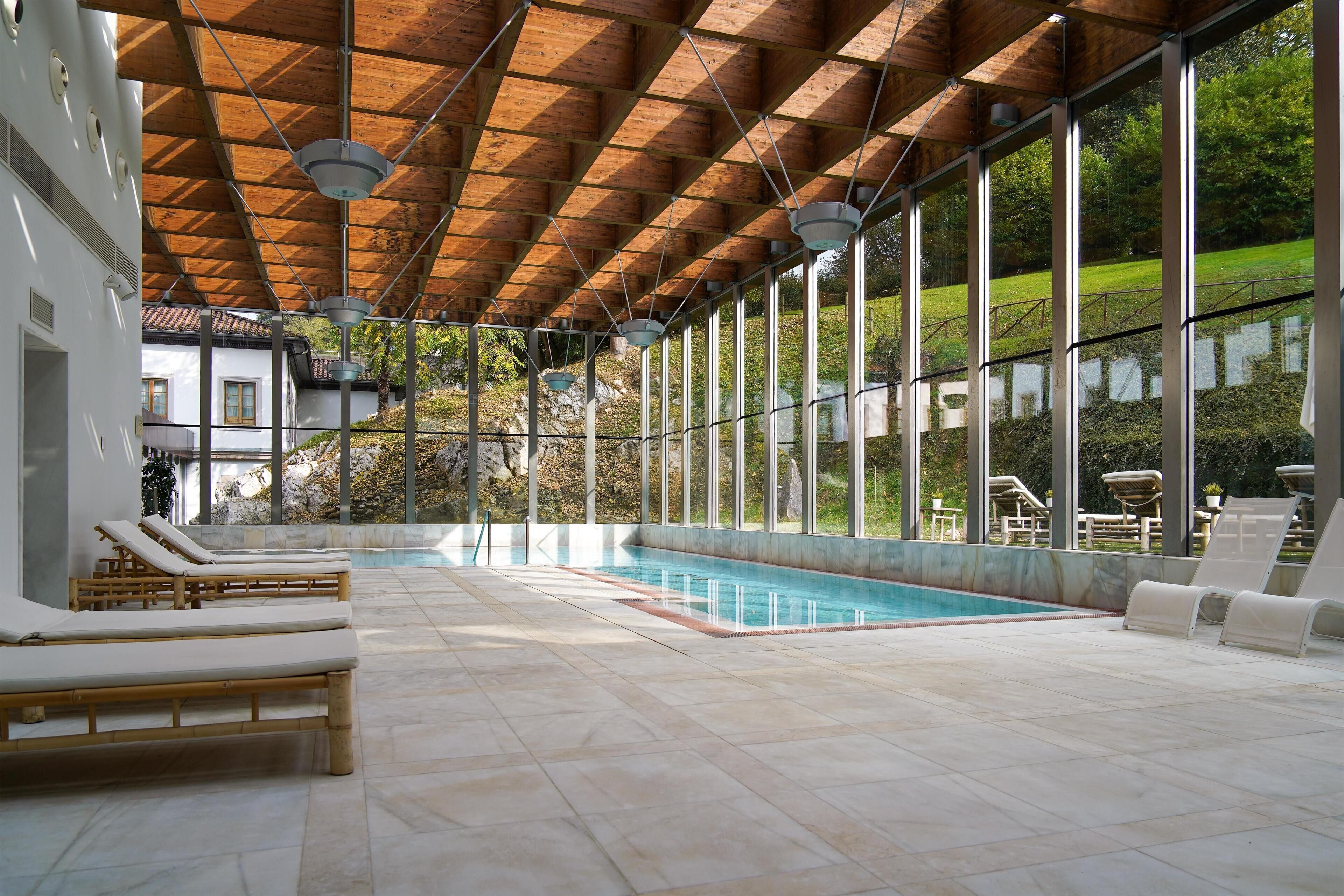 Una piscina interior moderna con vigas de madera en el techo, ventanas amplias y sillones de descanso.
