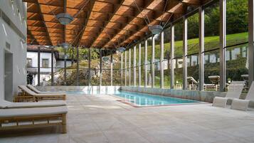 Una piscina interior moderna con vigas de madera en el techo, ventanas amplias y sillones de descanso.