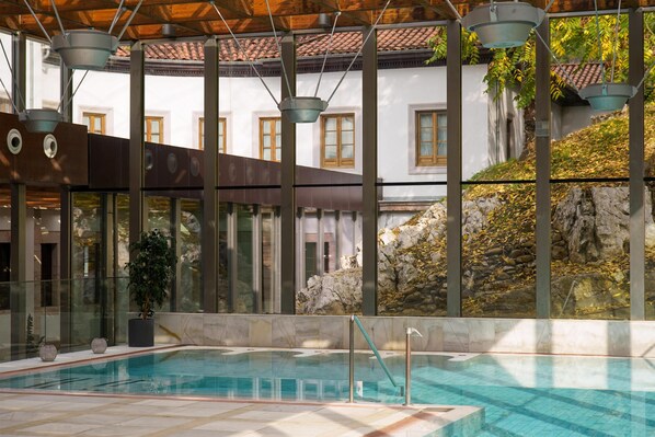 An indoor swimming pool with clear glass walls, a stone wall with a rocky outcrop, and a traditional building with wooden windows in the background.