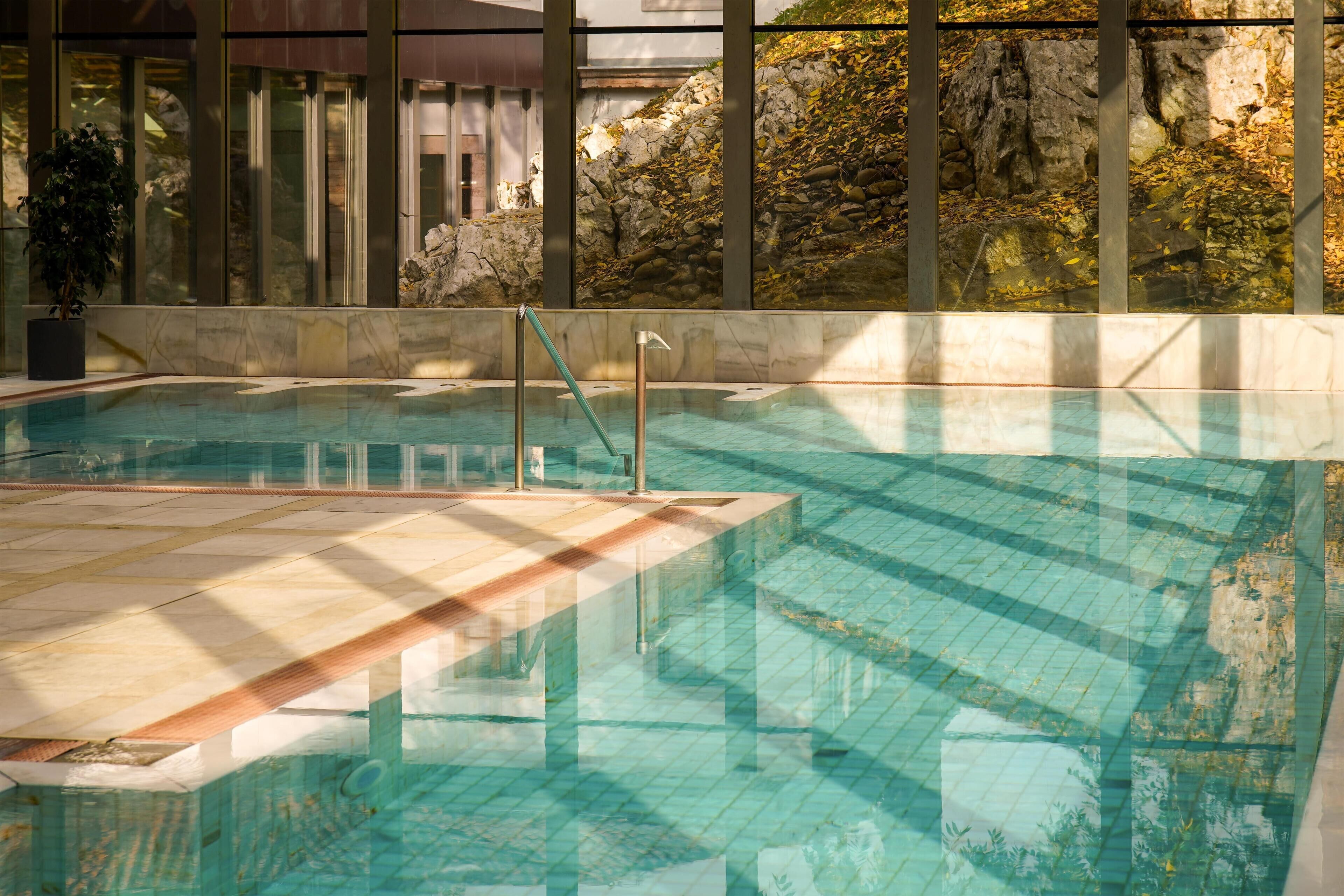 An indoor swimming pool with clear water, a tiled deck, and a view of rocky terrain through large windows.