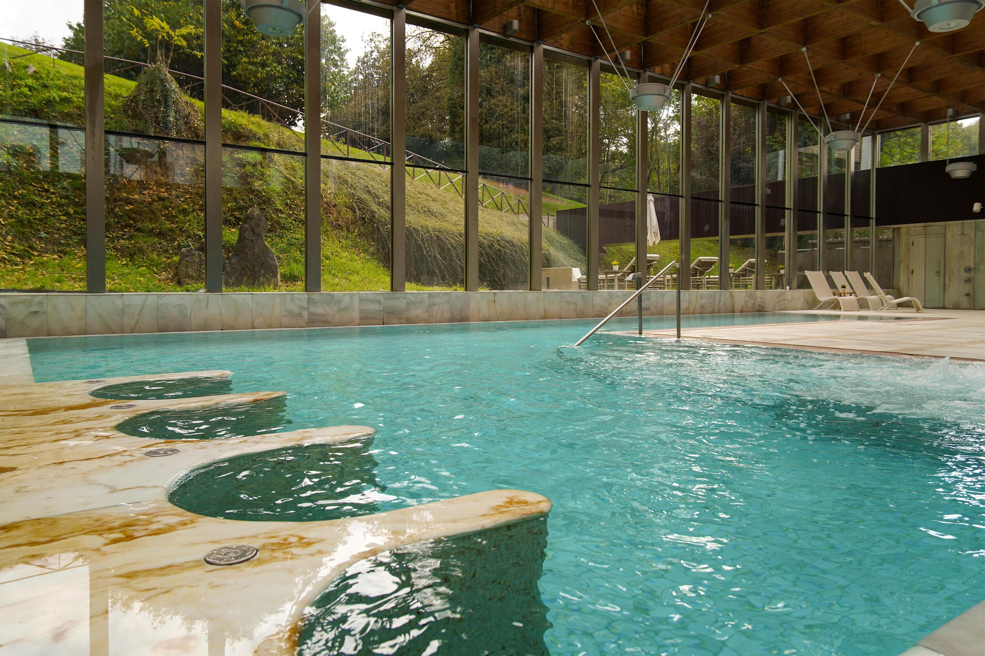 Una piscina interior con agua cristalina, techo de madera y vistas a una ladera cubierta de césped a través de amplios ventanales.