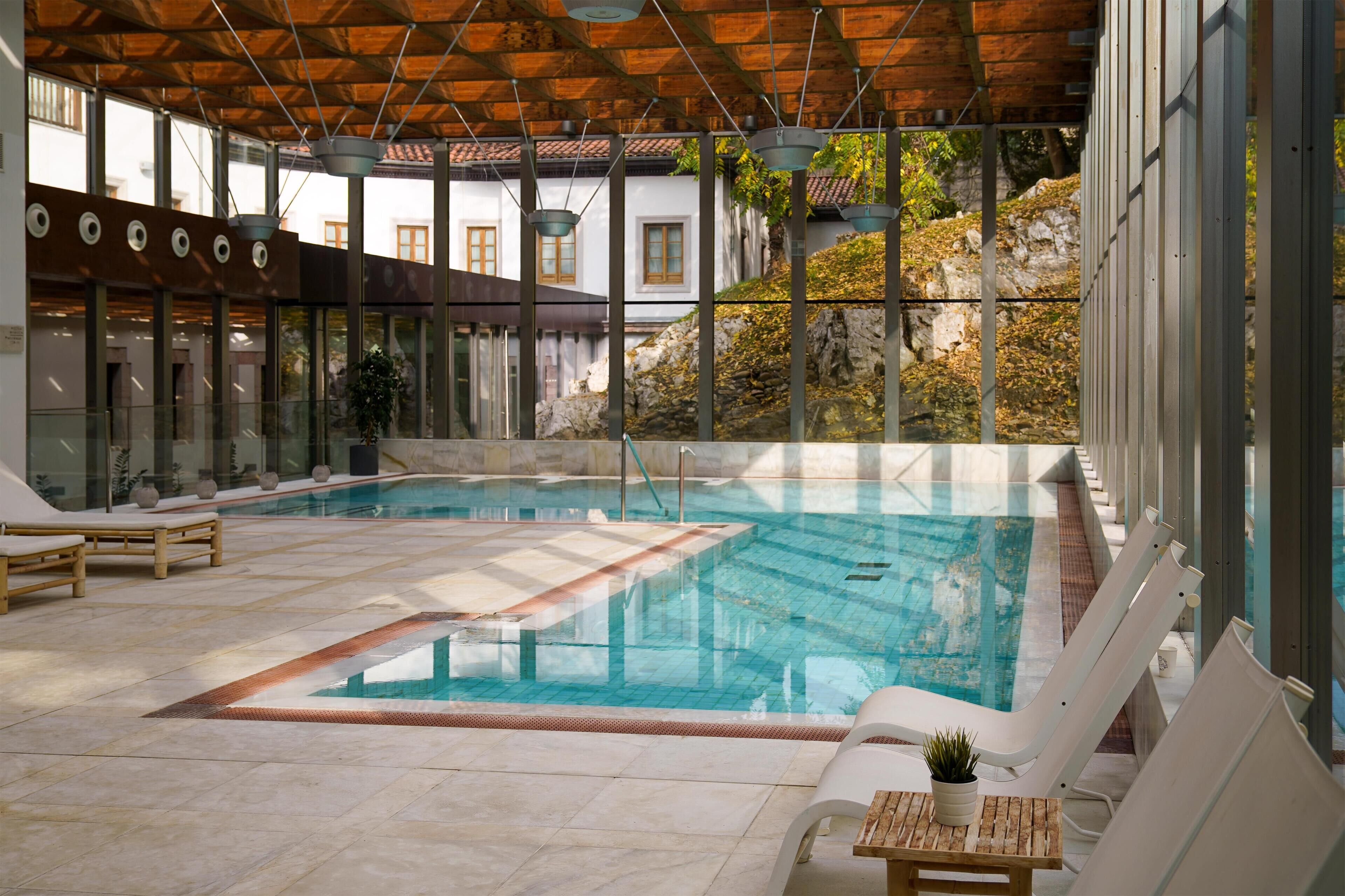 Una piscina interna con sedie a sdraio e un tavolo in legno, circondata da ampie finestre e vista su un paesaggio roccioso.