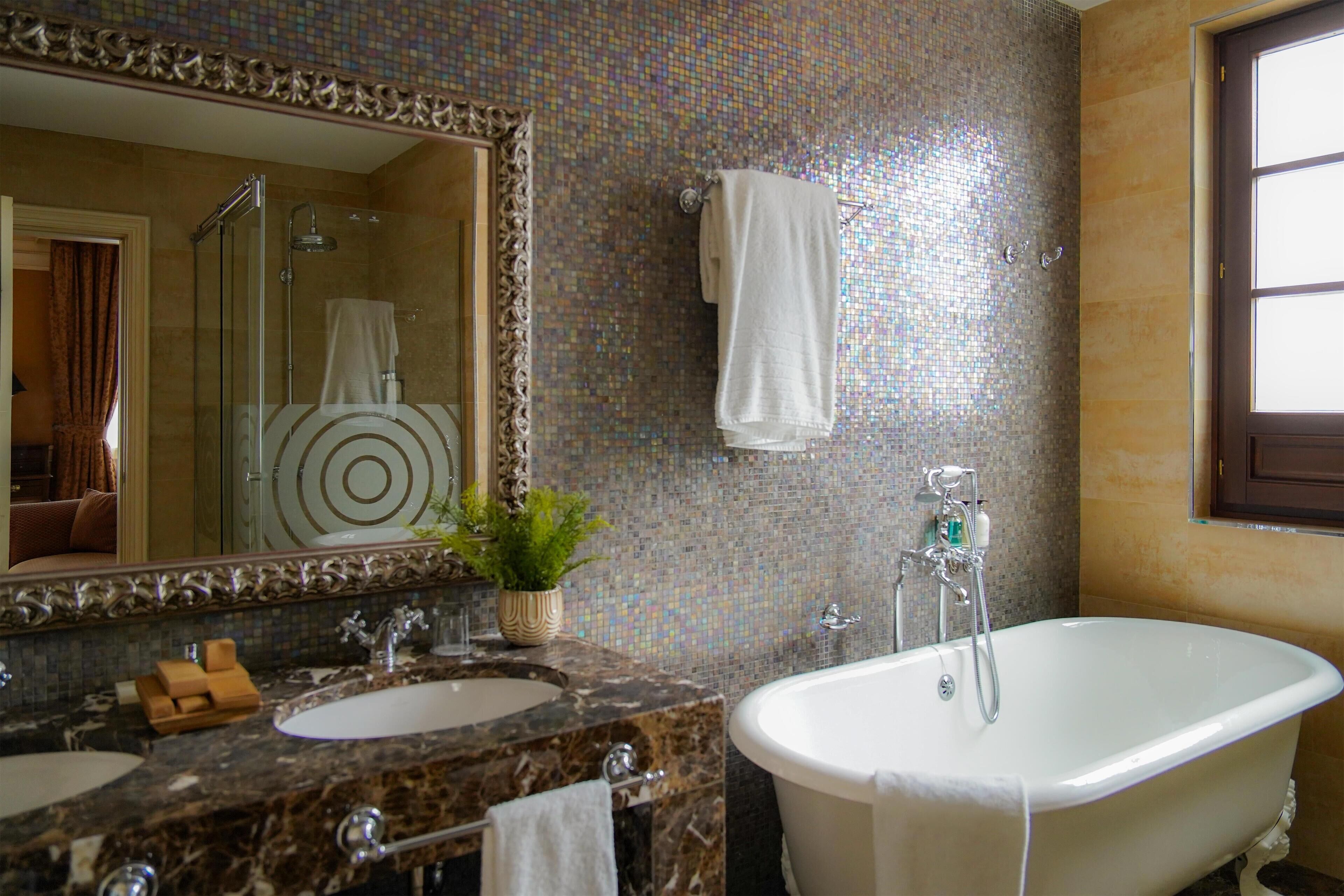A bathroom with a large mirror, two sinks, a bathtub, and a mosaic tile wall.