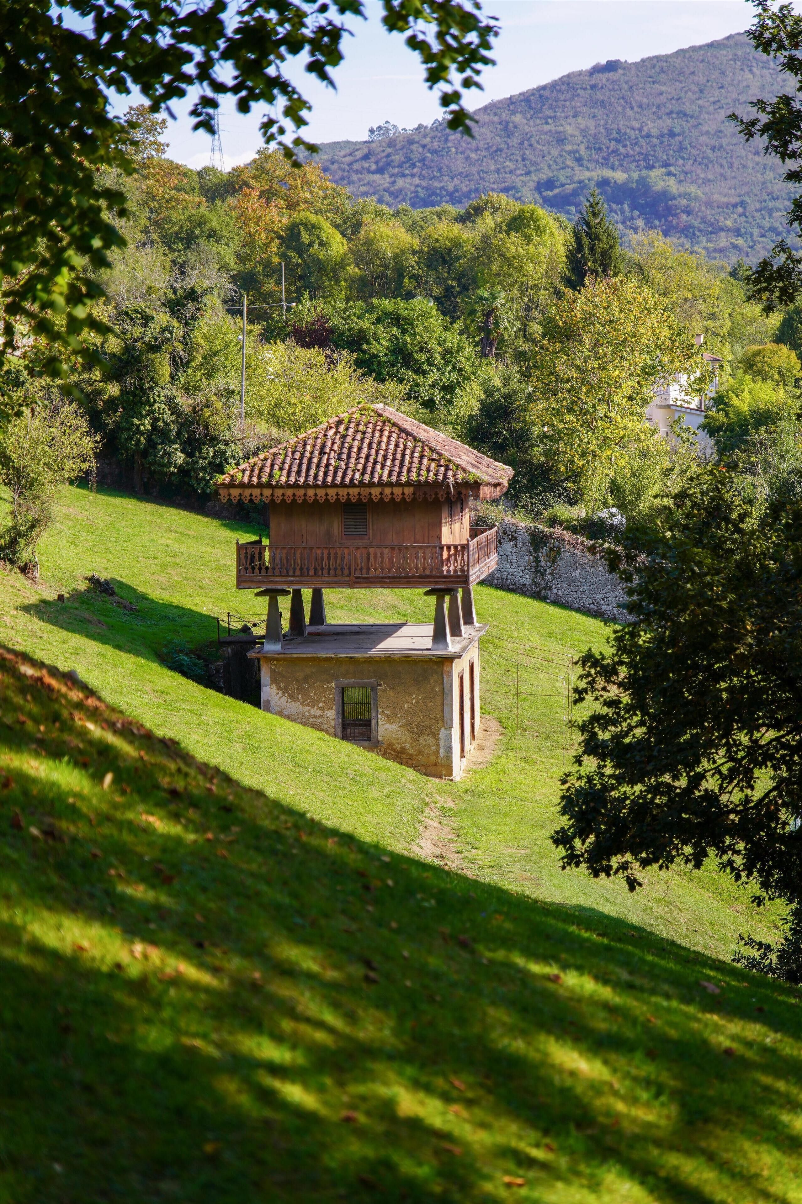 A wooden house with a balcony on a grassy hill, surrounded by trees and hills.