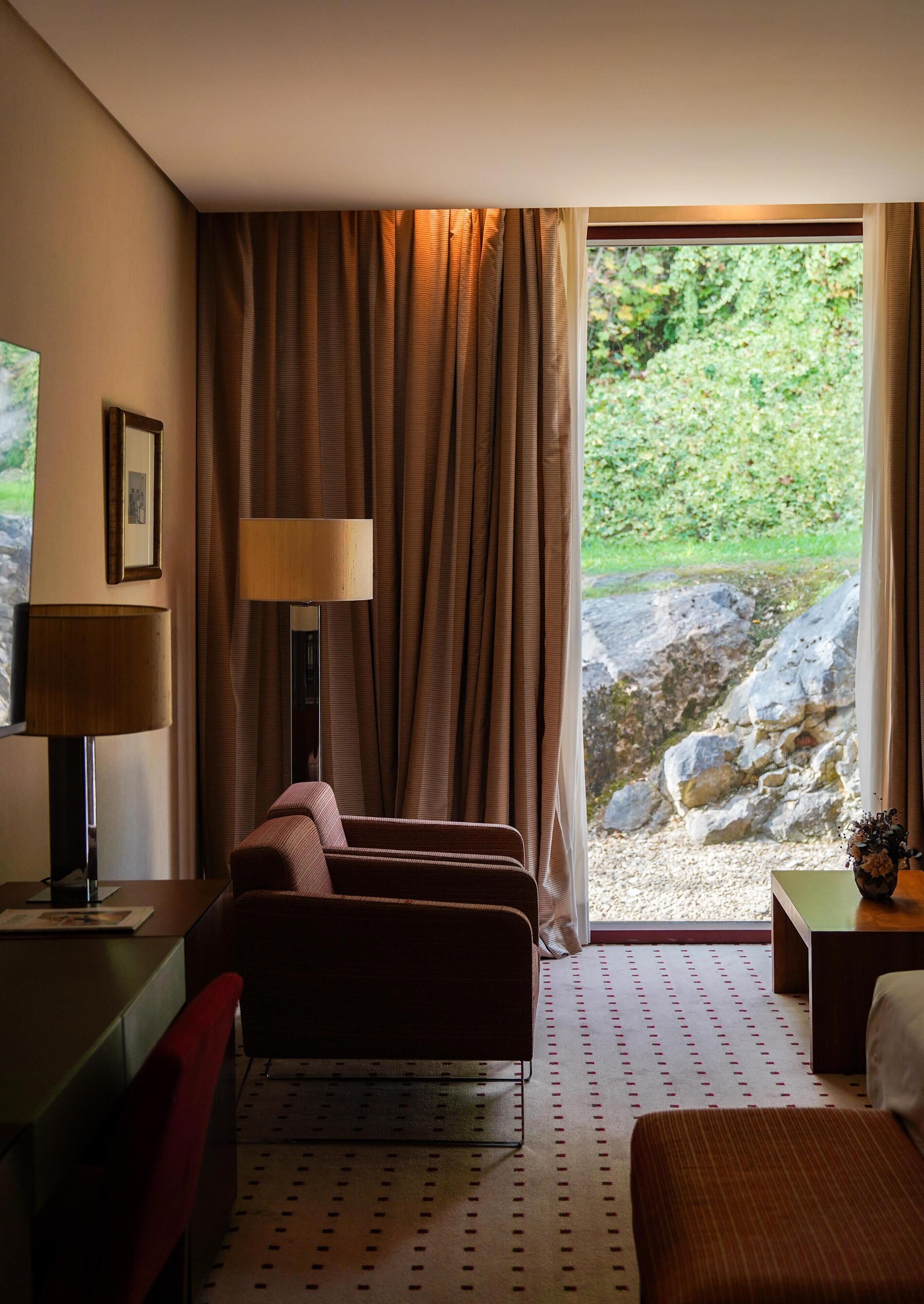 Habitación de hotel con cama, escritorio, silla, lámpara y vista a un área rocosa al aire libre.