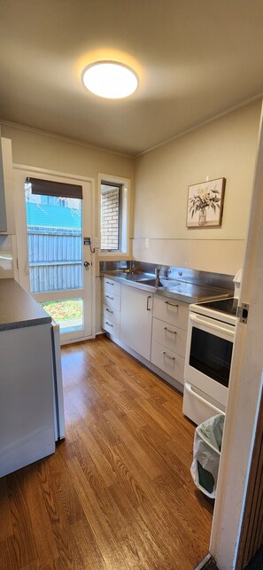 A compact kitchen with a refrigerator, oven, and sink, wooden flooring, a window with blinds, and a door leading outside. - Aalton Motel (Christchurch)