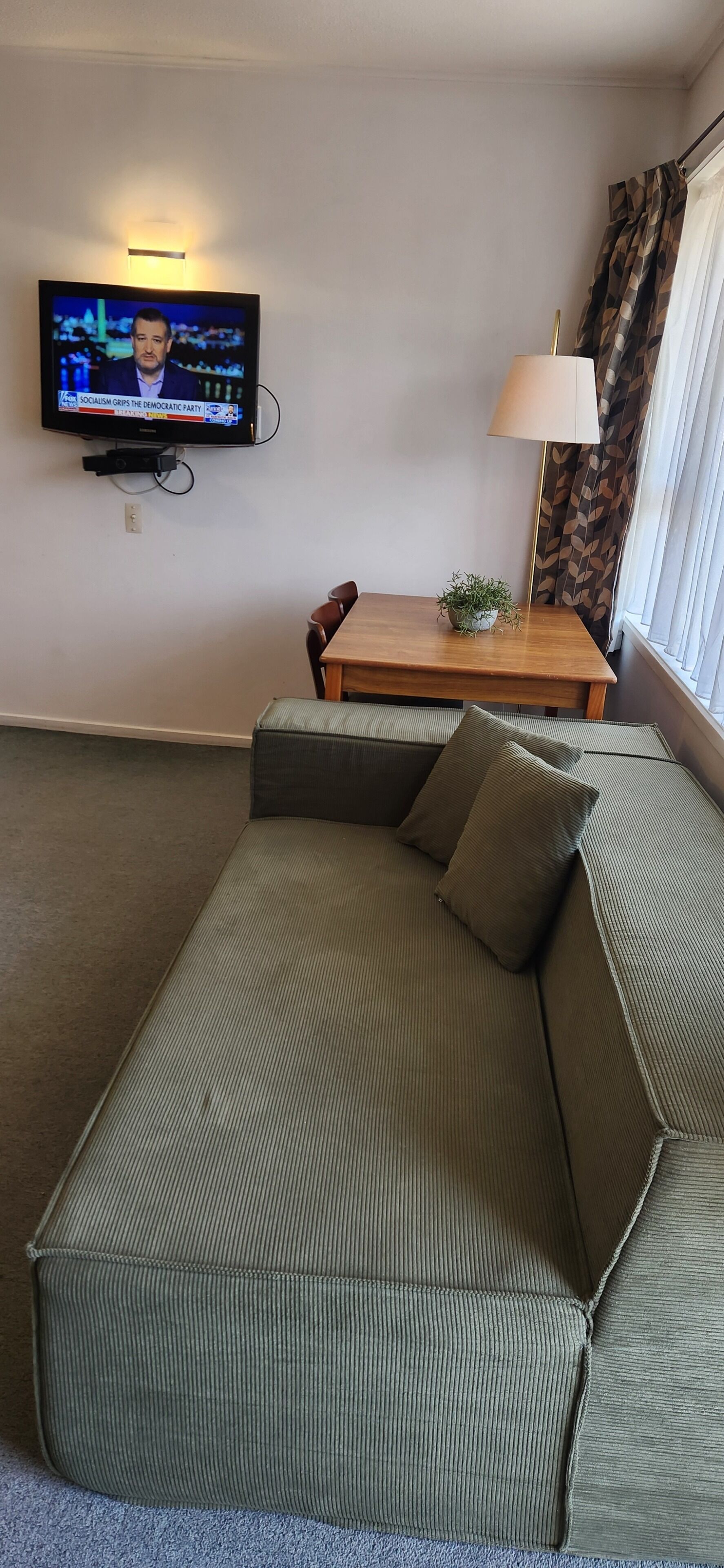 A living room with a green sofa, a wooden table with a lamp, and a television displaying a news channel.