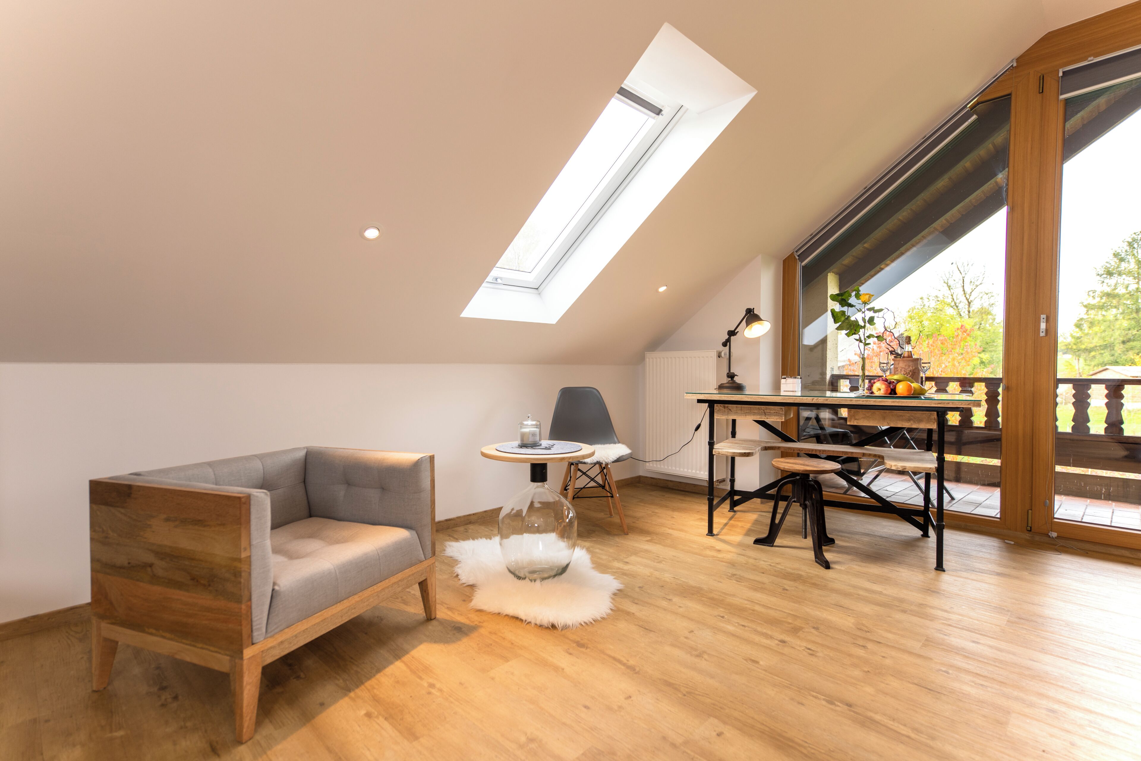 A loft-style room with a sloped ceiling, a sofa, a dining table with chairs, a small round table with a lamp, and a view through large windows.