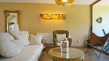 A living room with a sofa, a chair, a glass coffee table, a pendant light, a radiator, and a wall-mounted picture.