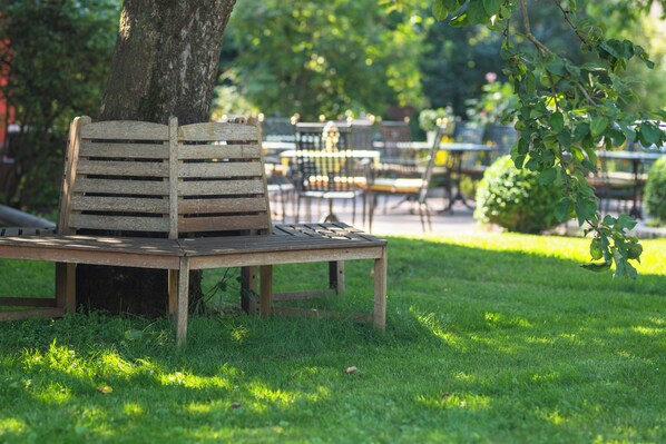 A wooden bench under a tree in a park with tables and chairs in the background. - Landhotel Lindenhof (Vohenstrauss)
