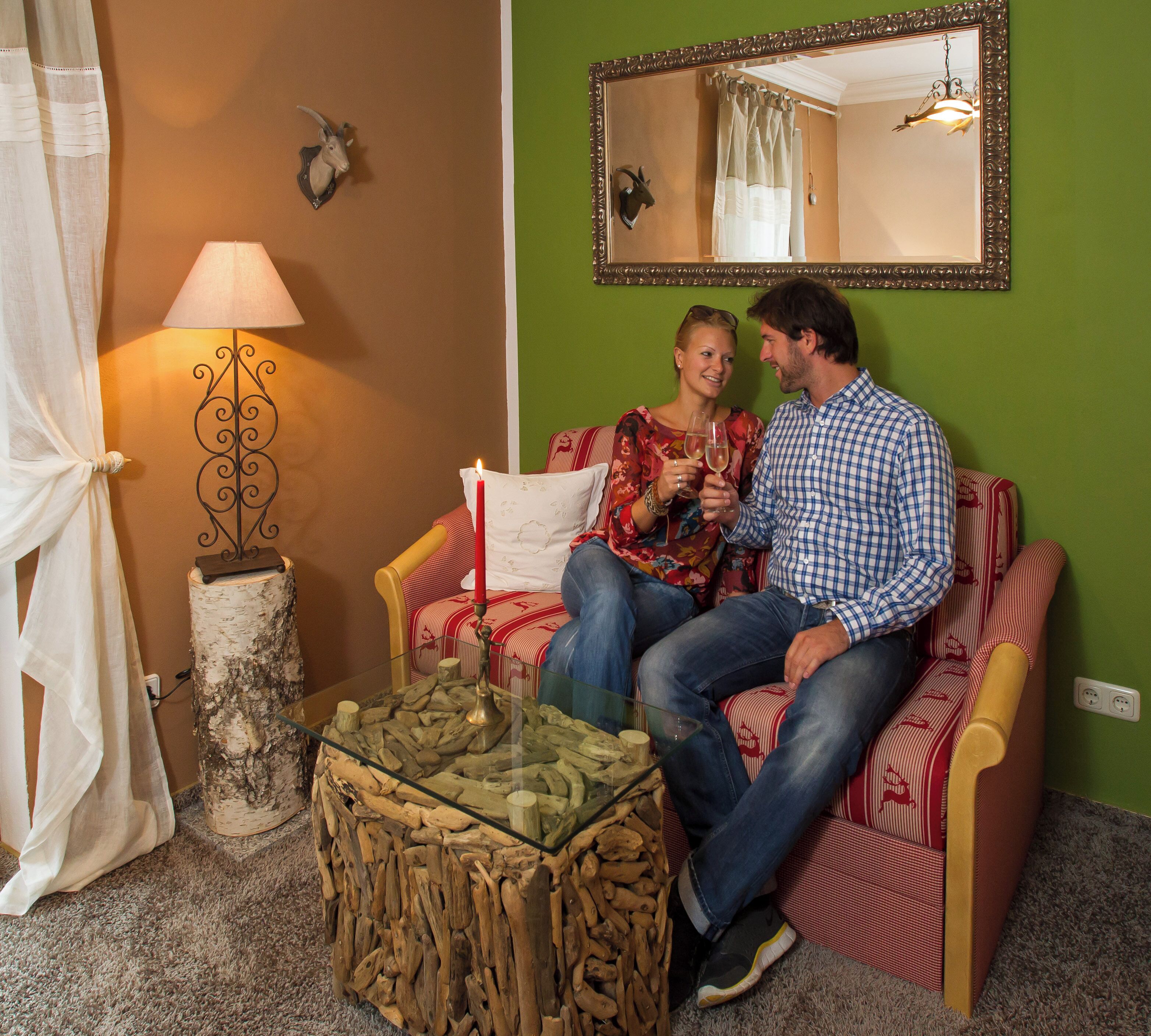 Een paar personen zitten op een rood-wit gestreepte bank, met een salontafel met houtblokken, een lamp, een spiegel aan de muur en een decoratieve dierenkop.