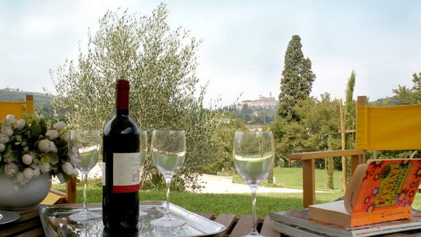 Una botella de vino y copas sobre una bandeja, un libro y un ramo de flores sobre una mesa al aire libre, con vistas a árboles y edificios.