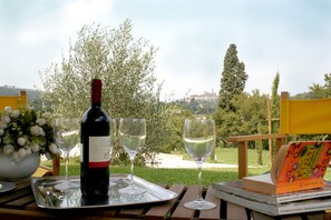 A wine bottle and glasses on a tray, a book, and a bouquet of flowers on a table outdoors with a view of trees and buildings.