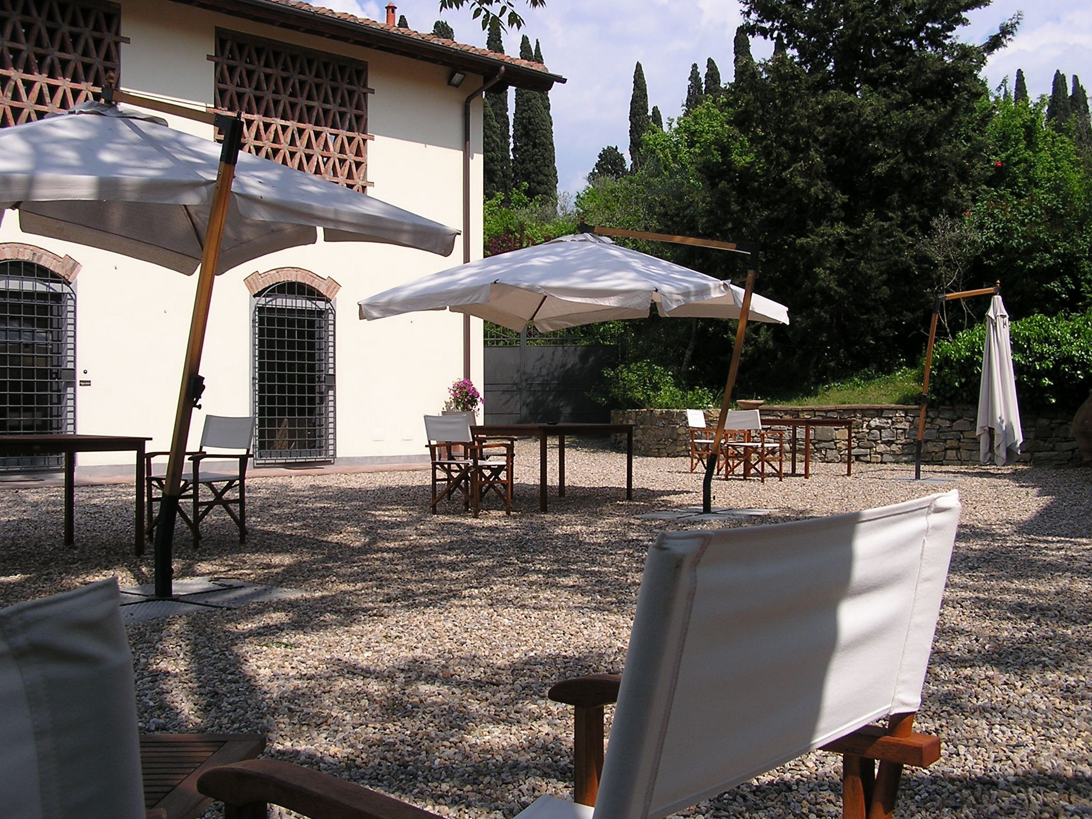 An outdoor seating area with tables and chairs, umbrellas, and a building with a tiled roof and arched windows.