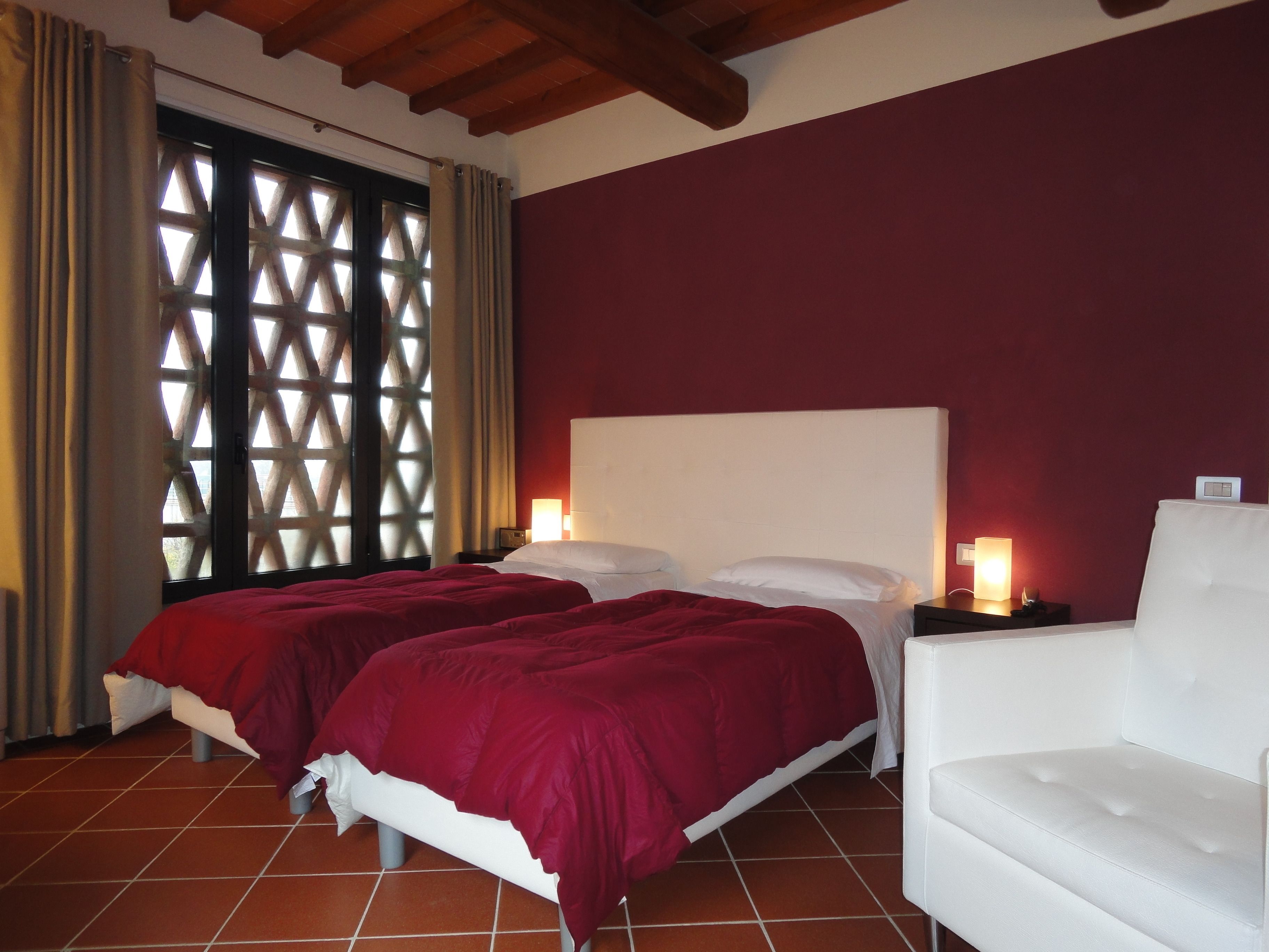 Habitación de hotel con dos camas, un sillón blanco, una ventana con diseño de celosía y una pared roja.