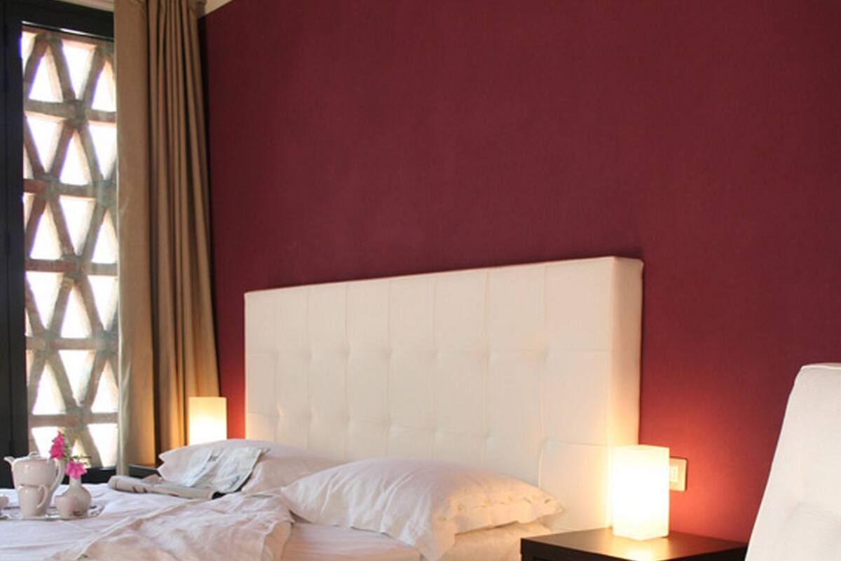 Un dormitorio con una cama grande, una mesita de noche con lámpara, una silla y una ventana con cortinas.