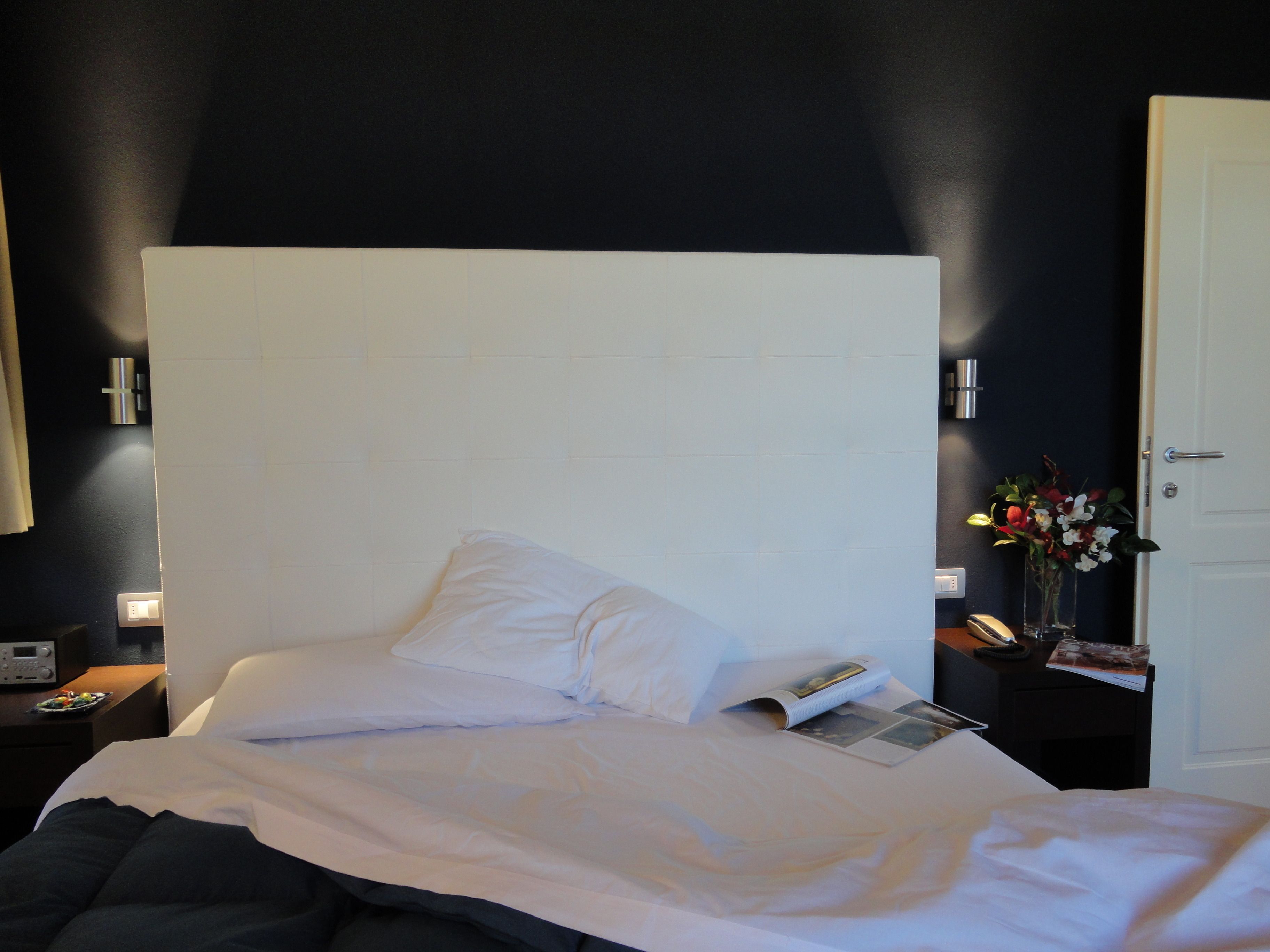A hotel room with a large bed, white bedding, a bedside table with a lamp, and a vase with flowers.