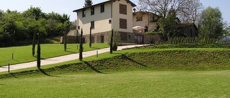 Un bâtiment à deux étages, doté d’une façade en pierre et d’un toit plat, entouré d’une pelouse bien entretenue et de quelques arbres.