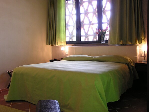 A hotel room with a bed, a window with curtains, a bedside table with a lamp, and a wall-mounted TV.